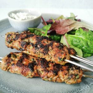 Lamb koftas on metal skewers with sald and a small bowl of mint yoghurt on a grey plate