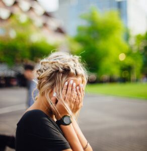 a blonde woman wit her hands covering her face in despair, sitting outside.