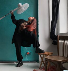 a woman in black, striking a pose, holding an electric fan to her face in front of a blue wall by a window