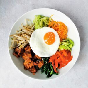 a white bowl on a mottled grey background with meat and various vegetables topped with a fried egg and sesame seeds