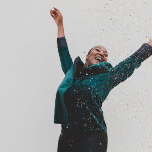 A woman in a green jacket with her arms up loving life with confetti raining down