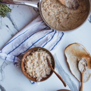 A bowl in a rustic bowl containing porridge and a steel pan with porridge on a white background with a blue striped tea towel