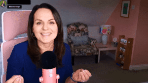 Natasha sitting in a pink office chair speaking into a pink blue yeti microphone with a floral chair and pink and white walls behind.