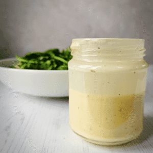 Lidless jar half filled with salad dressing and a white bowl of green salad leaves in the background