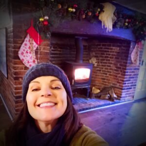 natasha grinning broadly with a wooly hat and scarf in front of a woodburner