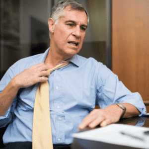 Man with grey hair, sitting at a desk with pen and paper, pulling at his tie because he is hot.